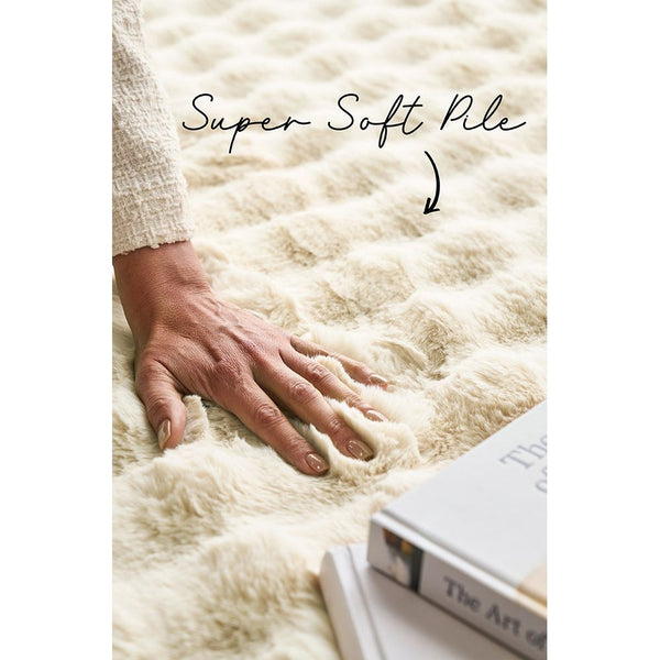 Close-up of soft, plush cream-colored rug with textured, wavy pattern and hand pressing into pile.