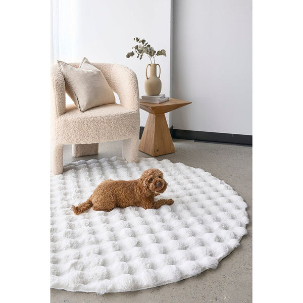 Round white rug with textured bubble pattern on gray floor next to beige upholstered chair and wooden side table.