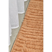 Close-up of a textured rust coloured rug on a concrete floor with a white curtain partially visible.