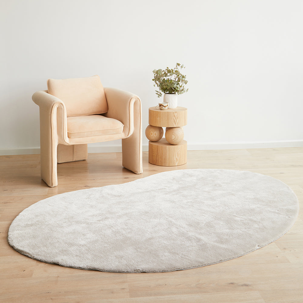 Oval pale beige soft rug on light wood floor next to beige modern cushioned chair and beige tiered side table with plant.