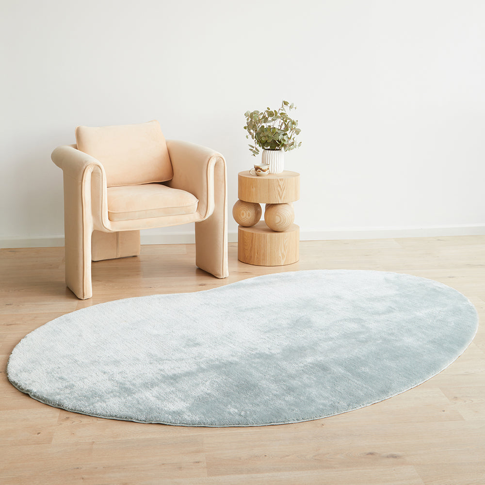Oval pale blue plush rug on light wood floor next to beige modern cushioned chair and round wooden side table with plant.