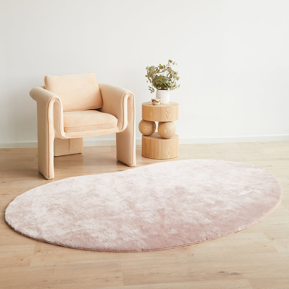 Oval pale pink plush rug on light wood floor beside beige modern armchair and round padded side table with plant.