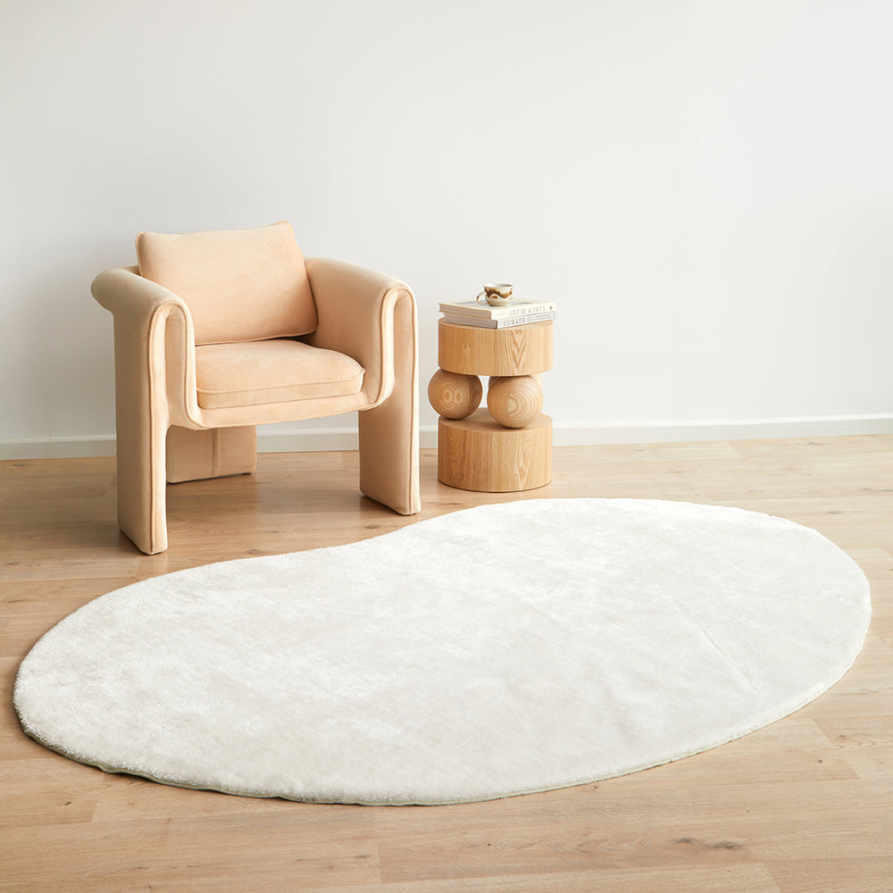 Oval white soft polyester rug on light wood floor beside beige cushioned armchair and wooden side table.