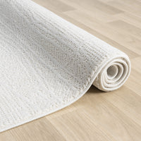 Cream-colored modern rug with subtle textured geometric pattern, partially rolled on light wood floor.
