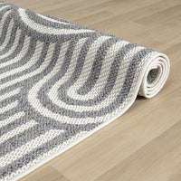 Grey flatweave rug with raised monochrome geometric arch pattern, rolled on wooden floor.