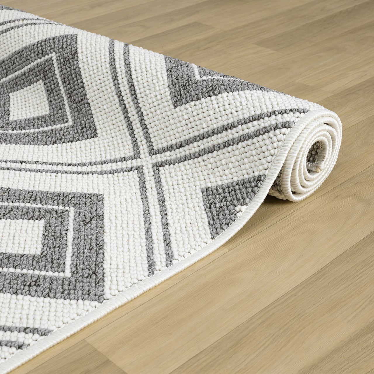 Rolled grey and white flatweave rug with bold geometric diamond pattern on light wood floor.