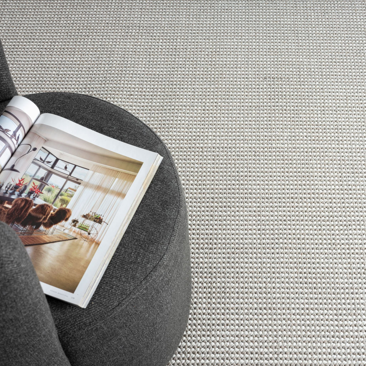 Grey flatweave rug with subtle geometric pattern next to dark grey fabric ottoman and open book.