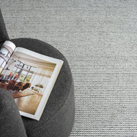 Grey flatweave rug with subtle textured pattern under dark grey ottoman and open magazine.