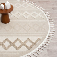 Beige round rug with cream geometric diamond patterns and fringe border on light wood floor.