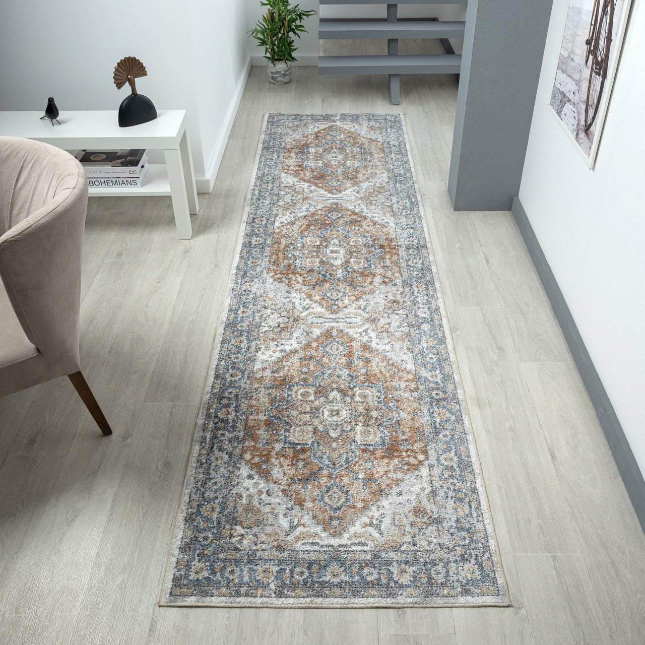 Long narrow runner rug with intricate geometric and floral patterns in blue, beige, and rust tones on a light background.