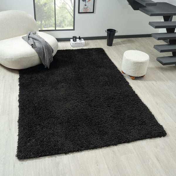 Black modern shaggy rug with dense polyester pile on light wood floor in contemporary living room setting.