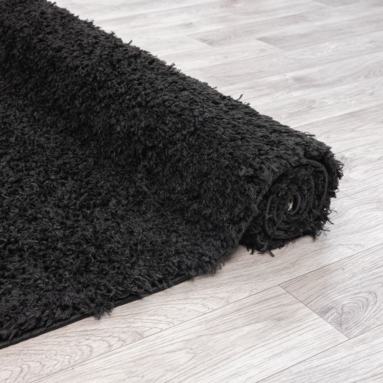 Black shaggy rug with thick polyester fibers, partially rolled on light wood floor.