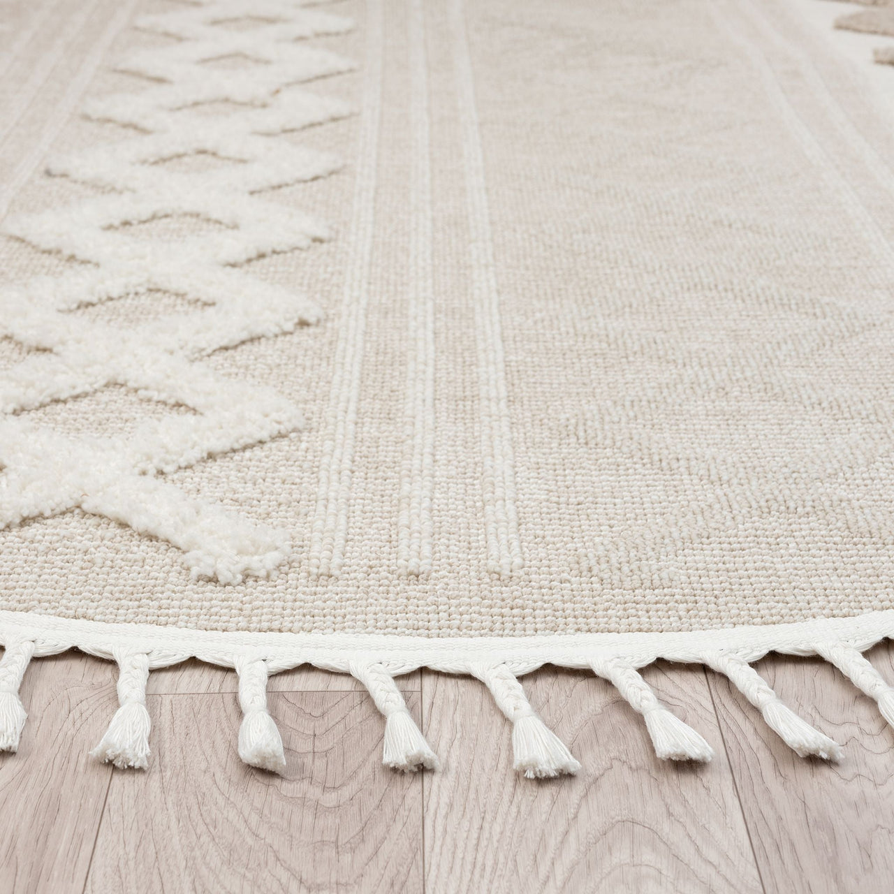 Beige and cream round rug with geometric textured pattern and fringe edges on light wood floor.