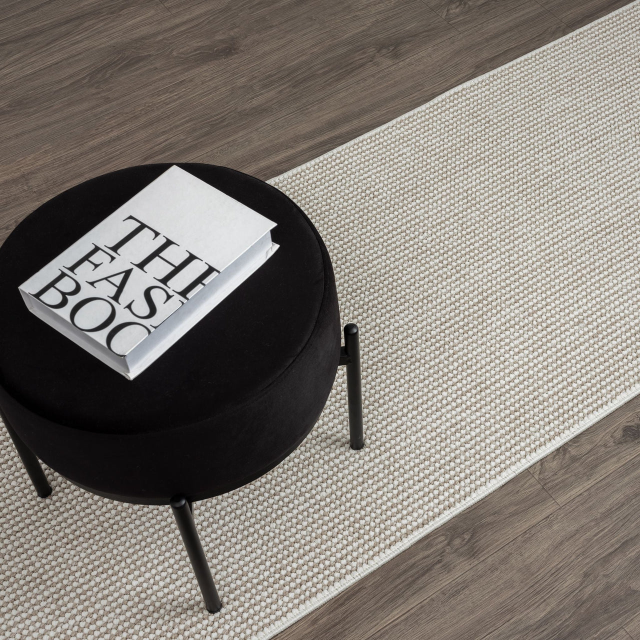 Beige flatweave runner rug with subtle geometric pattern on dark hardwood floor with black stool and book on top.