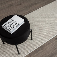 Beige flatweave runner rug with subtle geometric pattern on dark hardwood floor with black stool and book on top.