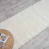 Cream shaggy runner rug on light wood flooring with a woven tray and magazines nearby.