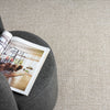 Taupe flatweave rug with subtle textured pattern beneath a dark gray round ottoman and an open magazine.