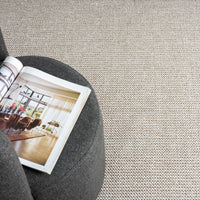 Taupe flatweave rug with subtle textured pattern beneath a dark gray round ottoman and an open magazine.