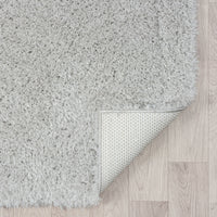 Grey modern shaggy rug with thick polyester fibers, square corner folded to show non-slip backing on light wood floor.