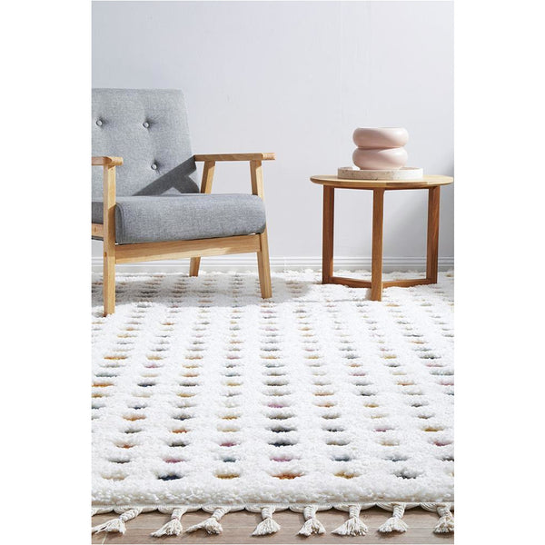 White shaggy rug with multi-colored polka dots and fringed edges under wooden chair and side table.