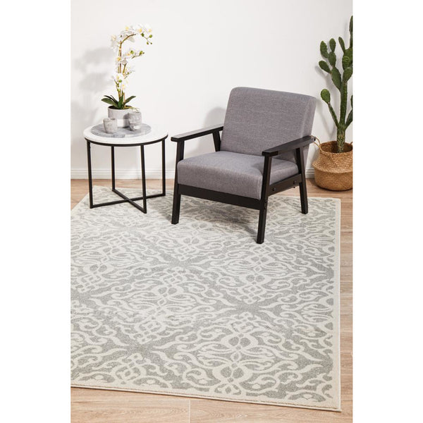 Silver grey and cream floral trellis patterned rug under a grey upholstered armchair with black frame and a round white side .