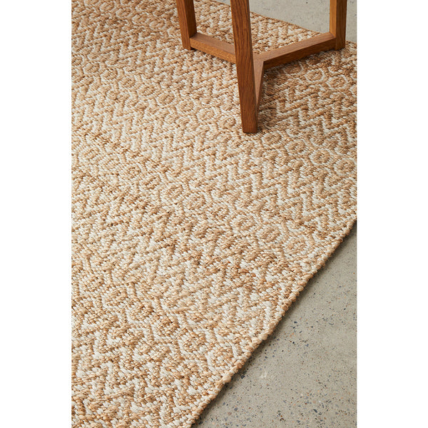 Natural jute and cotton flatwoven rug with geometric pattern under wooden chair leg on gray floor.