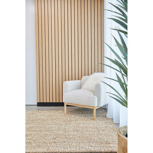Natural jute rug with chunky hand-loomed texture in a warm beige tone beneath a modern white armchair and wooden panel wall.
