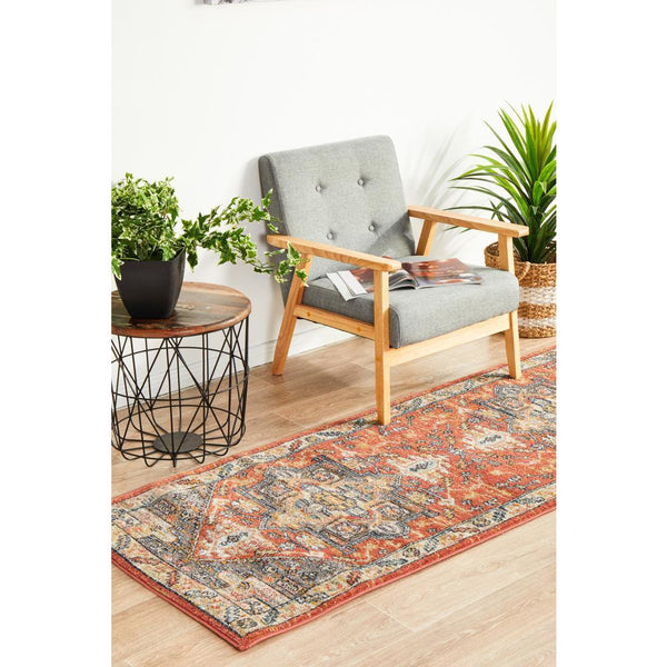 Terracotta and multicolor tribal patterned runner rug on light wood floor next to gray cushioned wooden chair and plants.