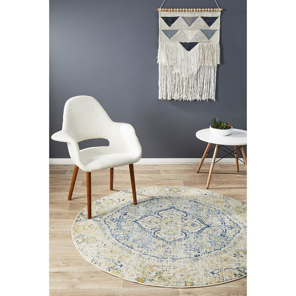 Round blue and beige patterned transitional bohemian rug on wooden floor by white chair and modern side table.